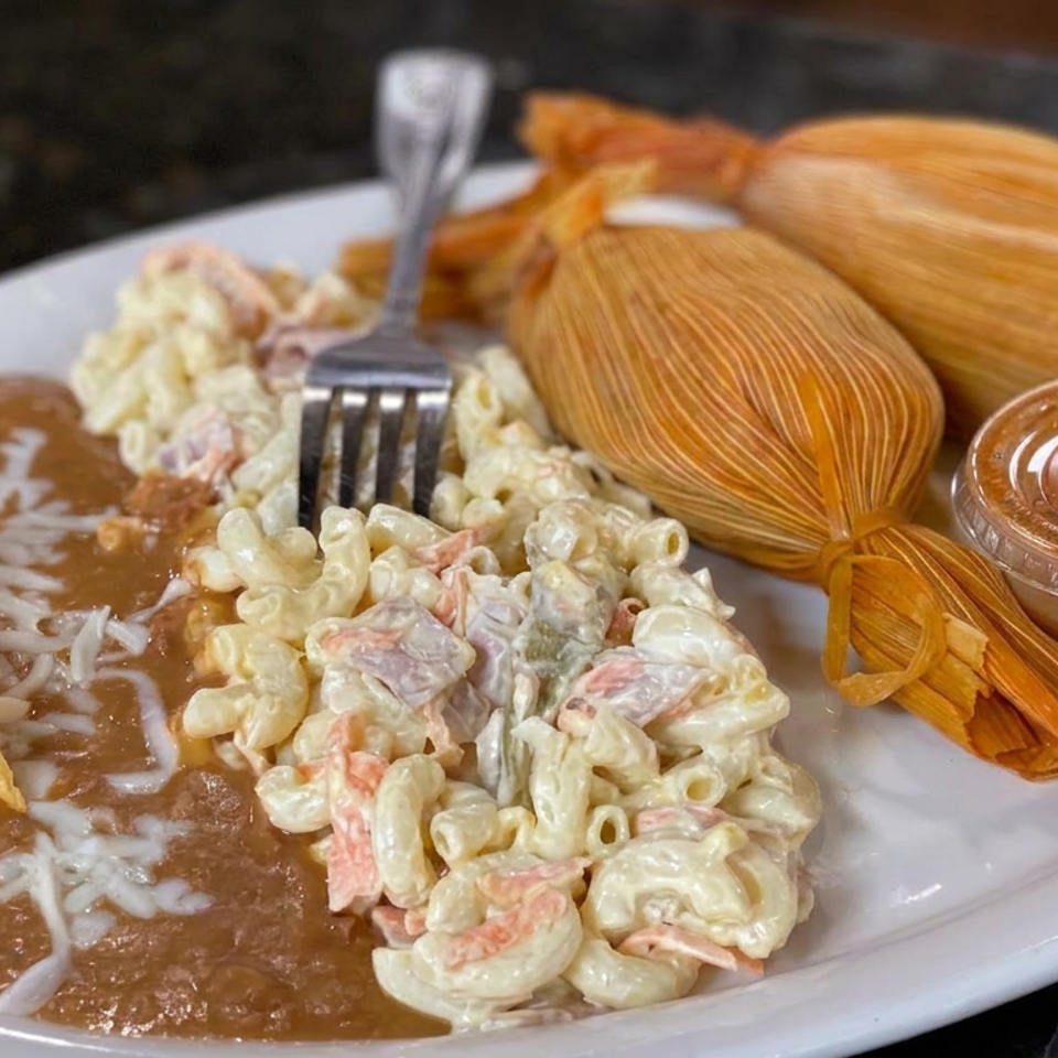 Plate with refried beans, rice, creamy macaroni salad, wrapped tamales, tortilla chips, and salsa.
